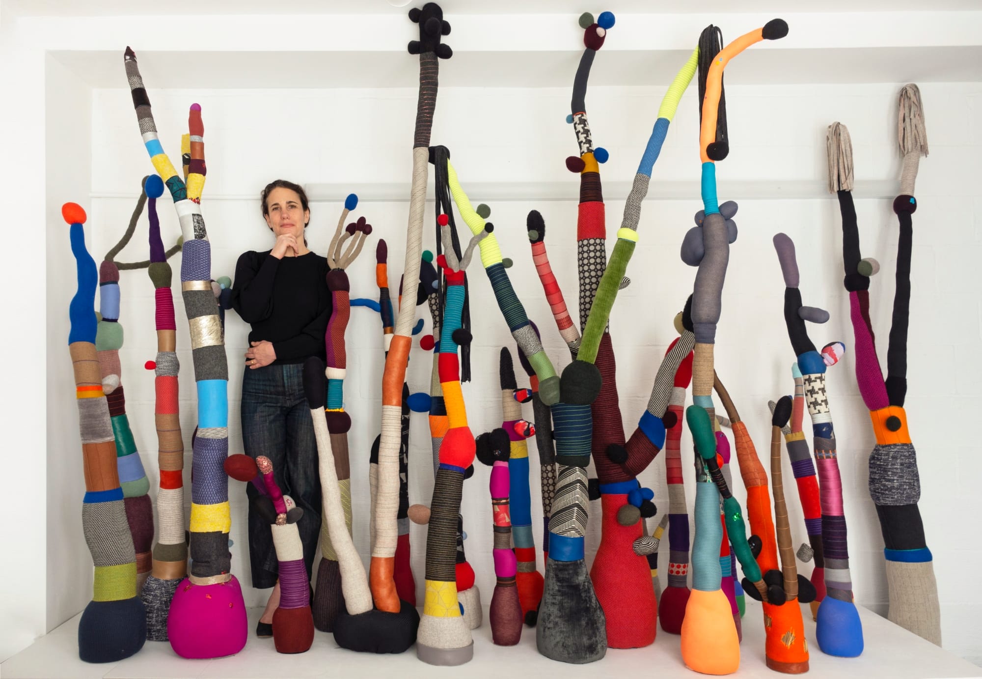 a white woman in jeans and a black top stands amid a cluster of vibrant textile structures
