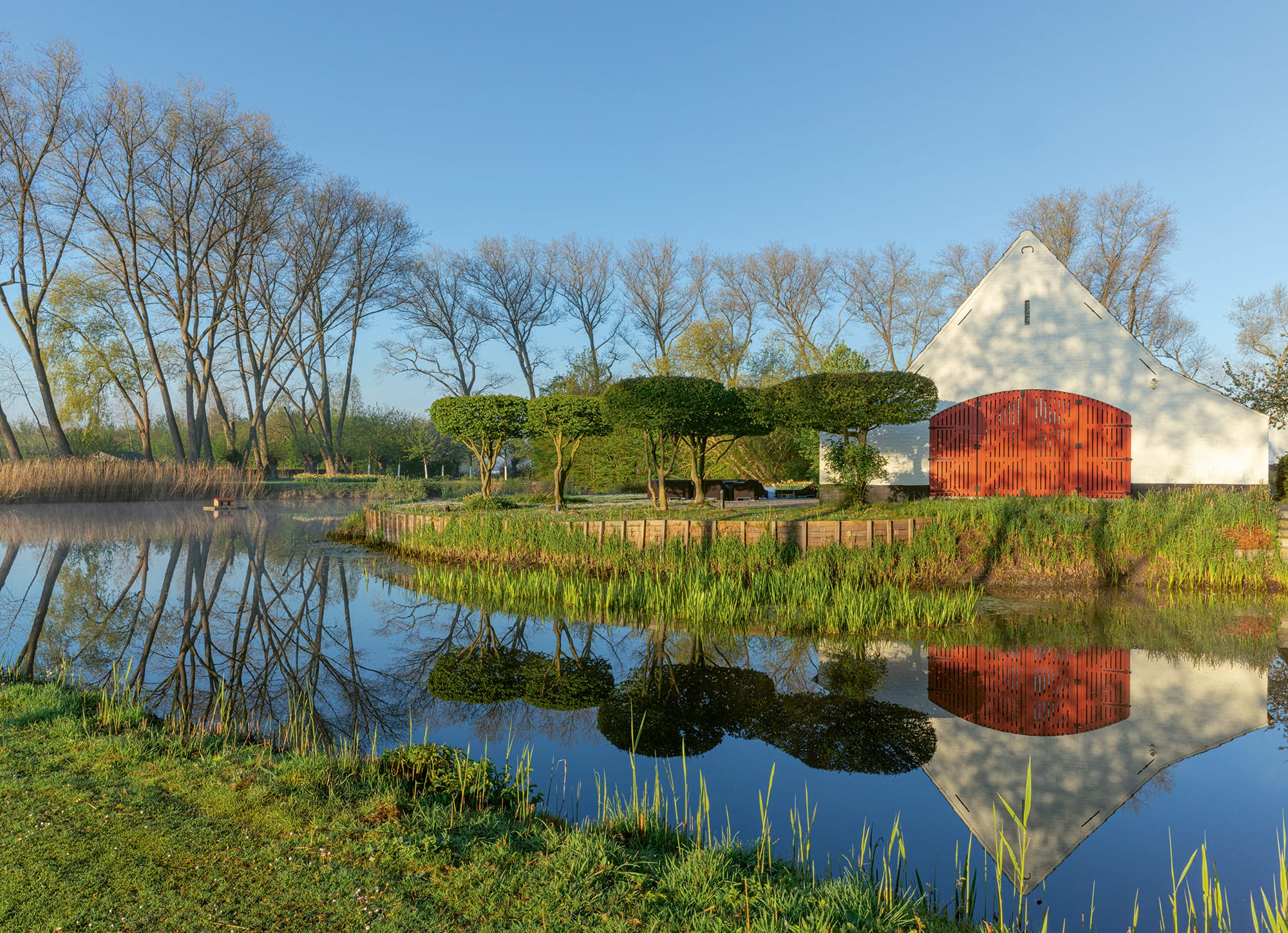 a white building with circular trimmed trees in the background with a stream flowing in the foreground